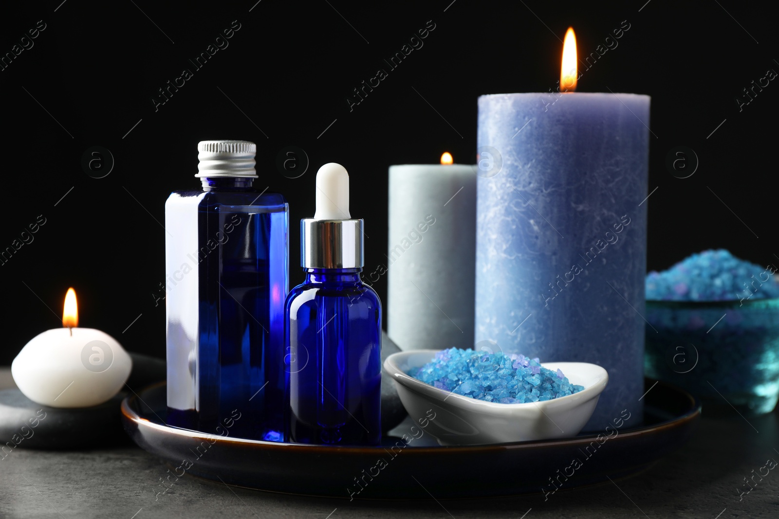 Photo of Spa composition. Cosmetic products, burning candles and sea salt on gray table against black background, closeup