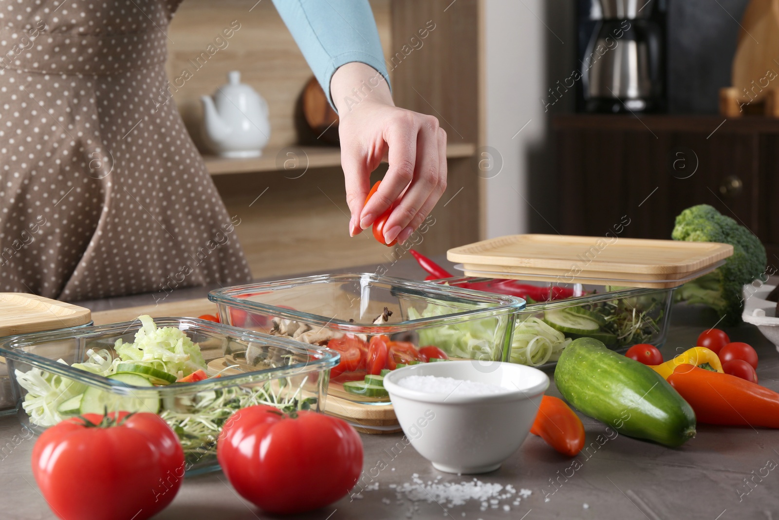 Photo of Healthy food. Woman putting piece of tomato into glass container at grey table in kitchen, closeup