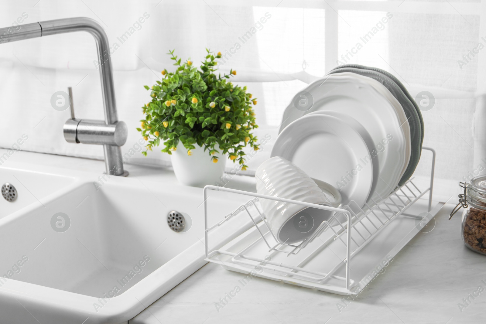 Photo of Drainer with different clean dishware, cup and houseplant near sink on light table indoors