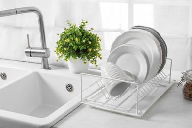 Drainer with different clean dishware, cup and houseplant near sink on light table indoors