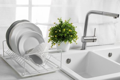 Photo of Drainer with different clean dishware, cup and houseplant near sink on light table indoors