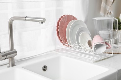 Drainer with different clean dishware and cup on light table near sink indoors