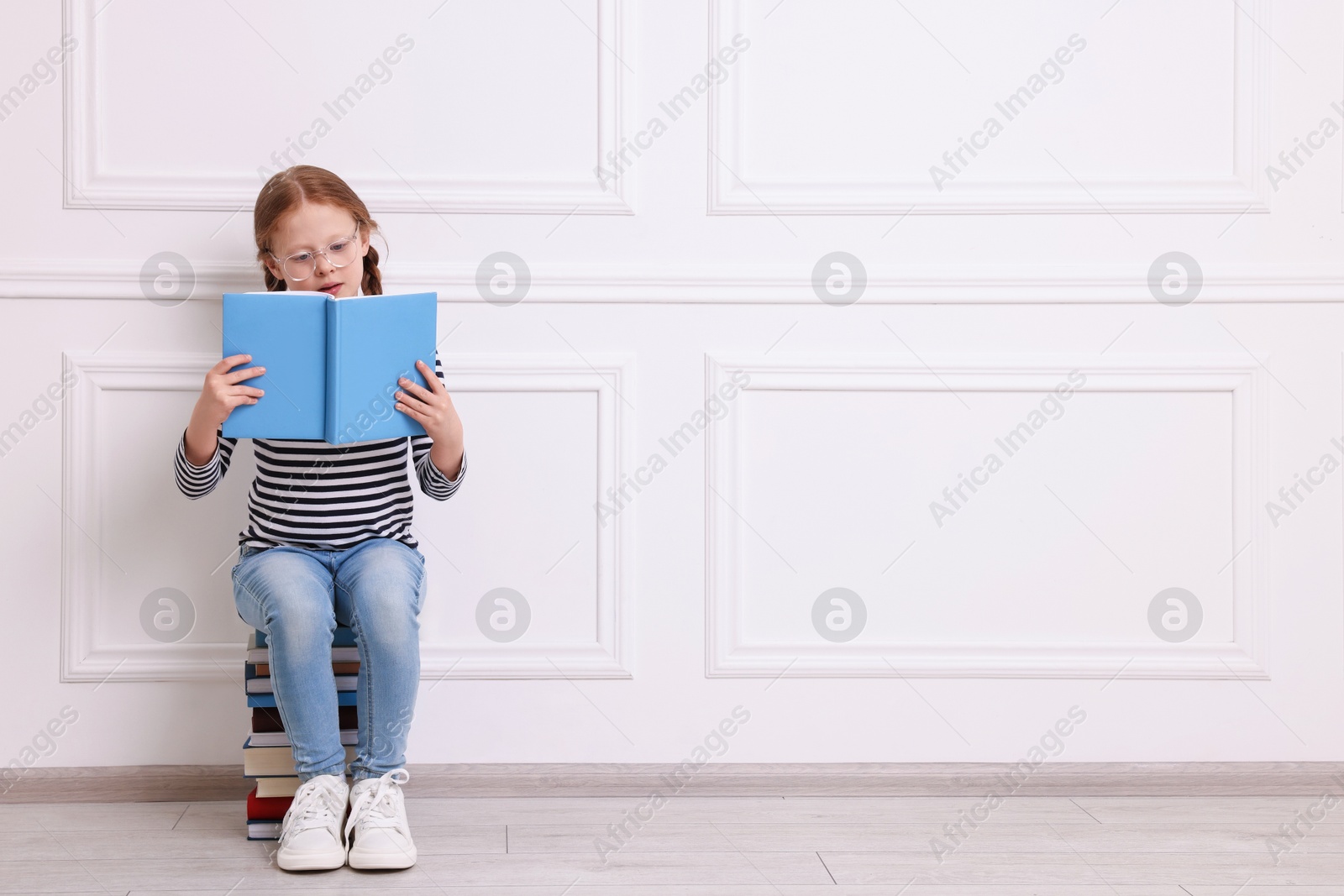 Photo of Cute girl reading while sitting on stack of books indoors. Space for text