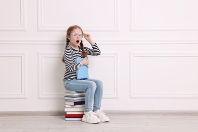 Surprised girl sitting on stack of books indoors. Space for text