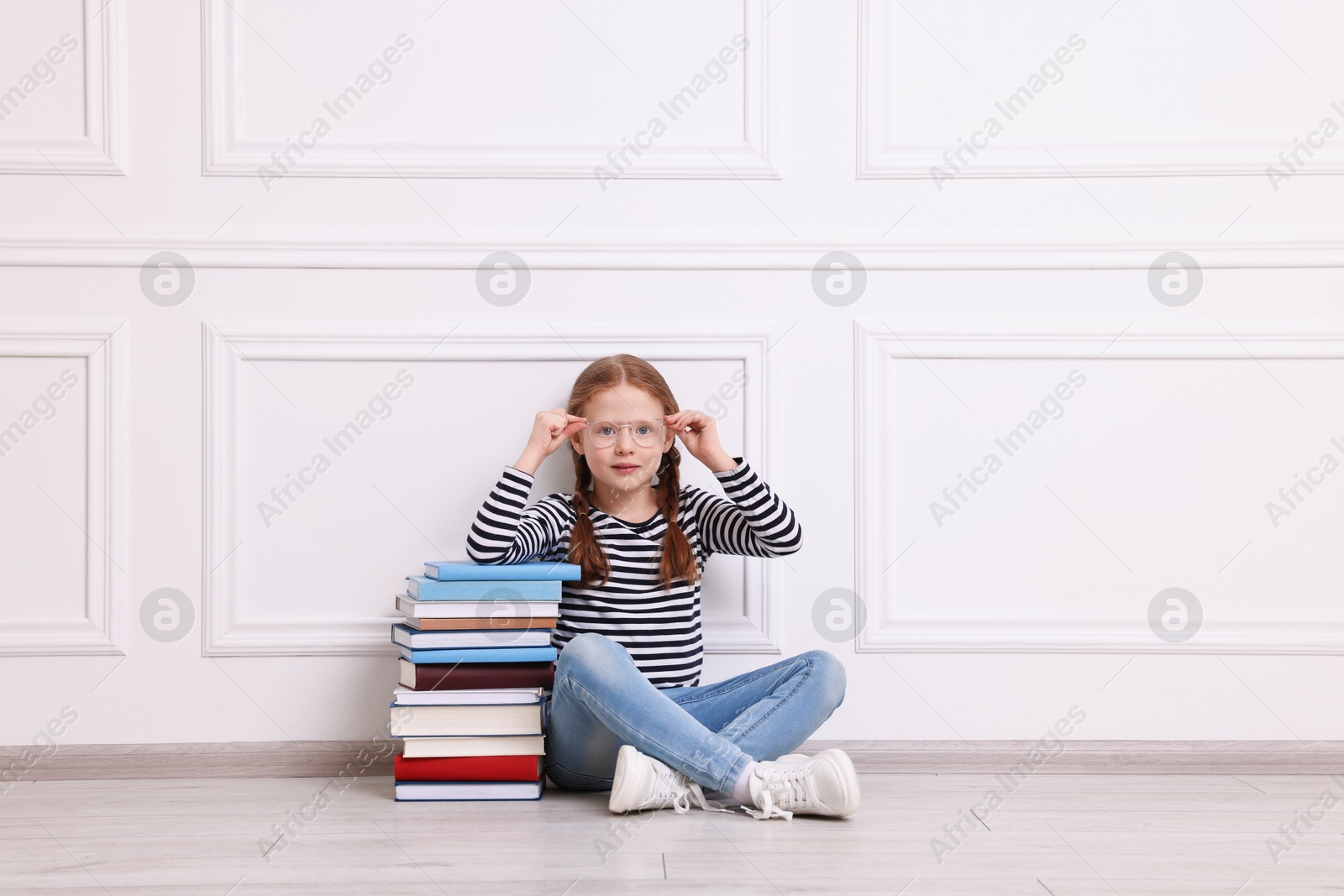 Photo of Cute girl sitting with stack of books indoors