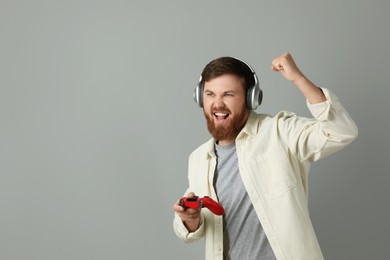 Emotional man in headphones with game controller on grey background. Space for text