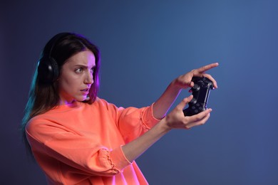 Woman in headphones playing video game with controller on dark blue background. Space for text