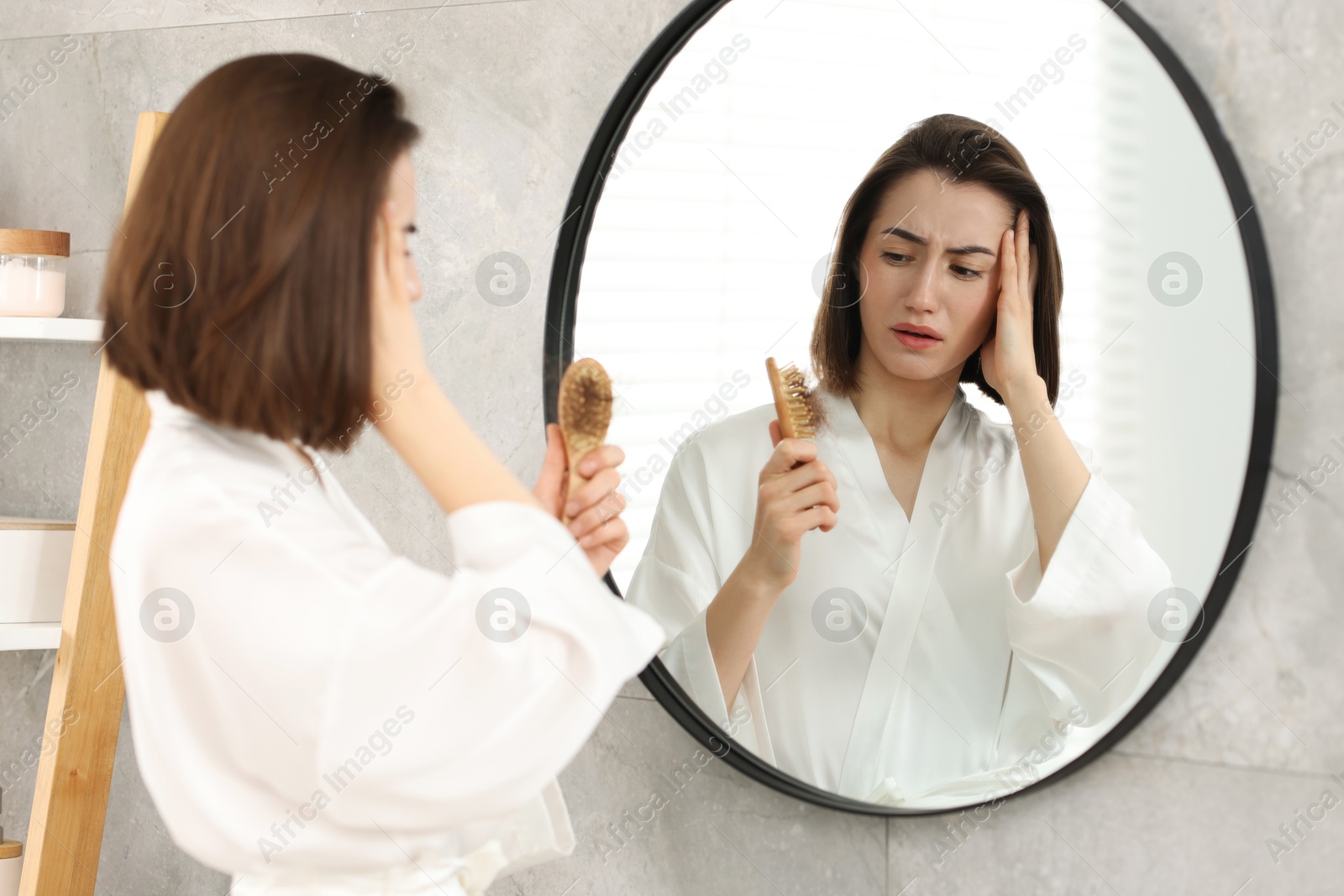 Photo of Stressed woman holding brush with lost hair near mirror at home. Alopecia problem