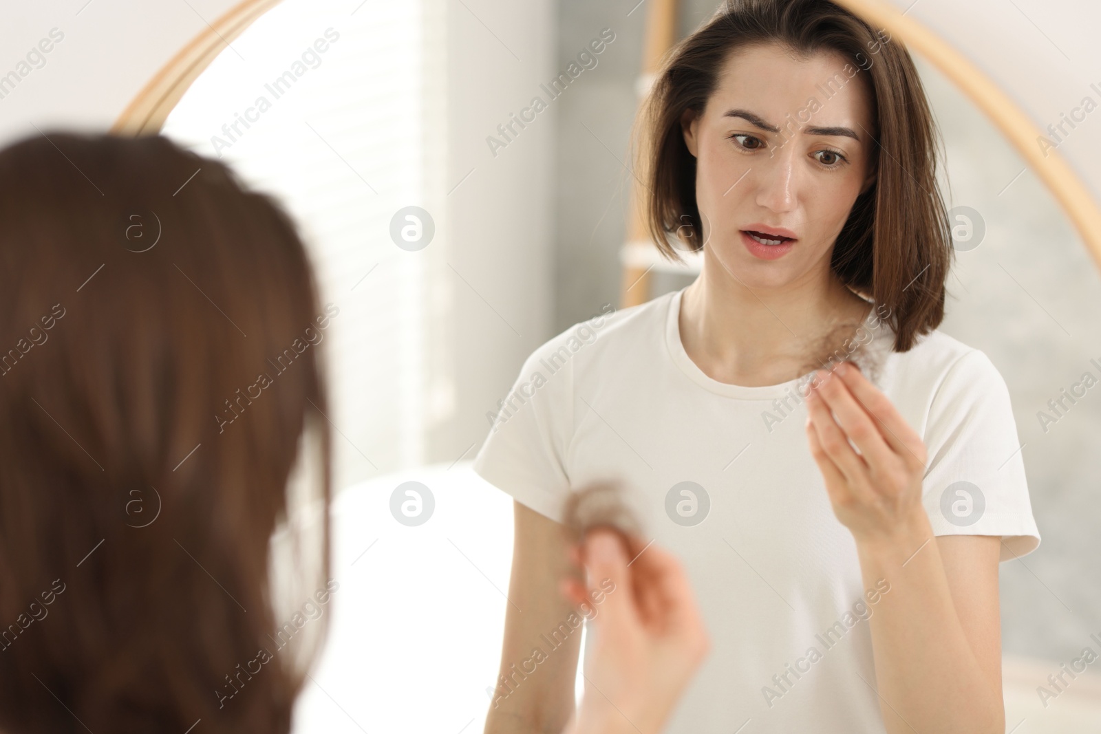 Photo of Stressed woman holding clump of lost hair near mirror indoors. Alopecia problem