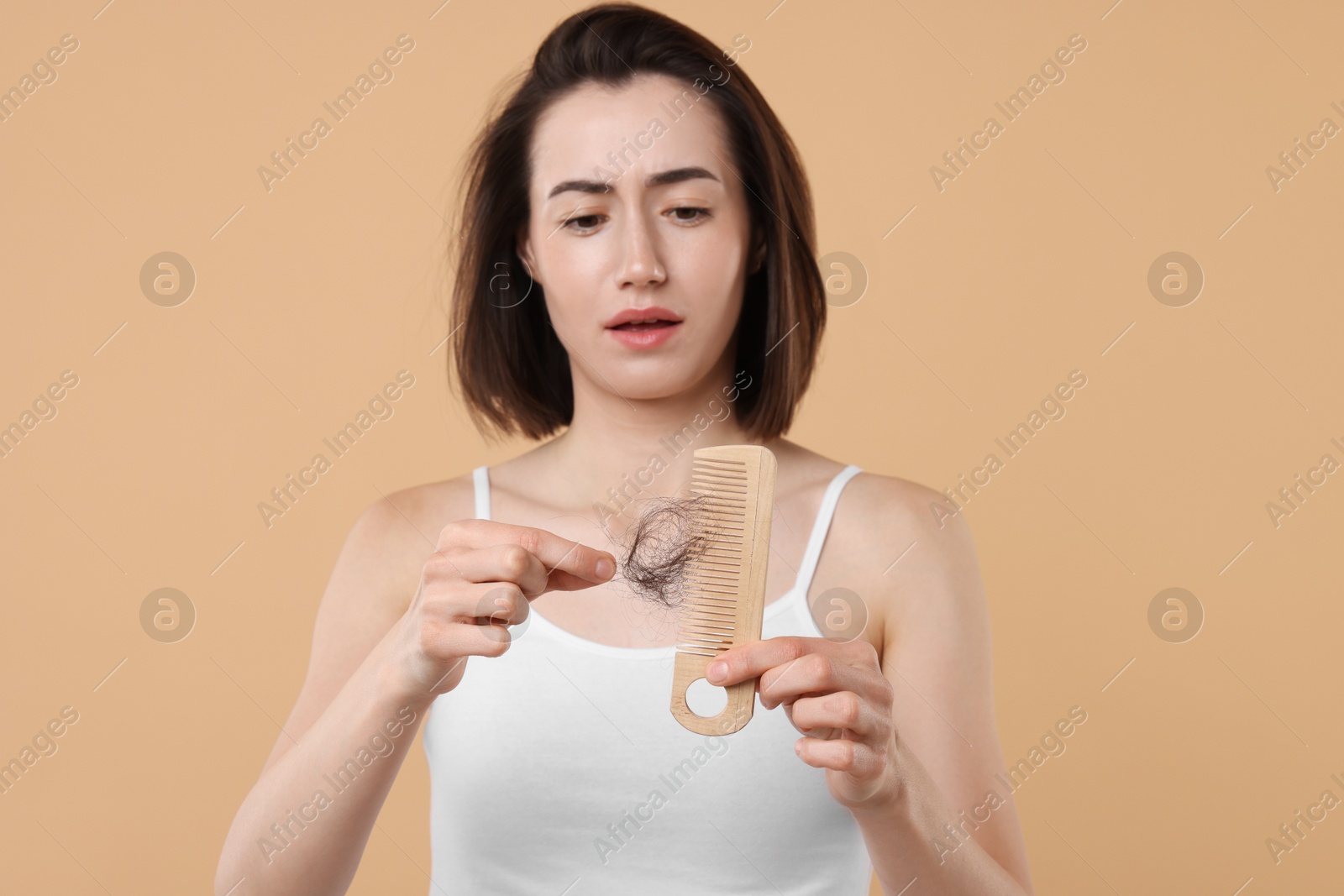 Photo of Sad woman taking her lost hair from comb on light brown background. Alopecia problem