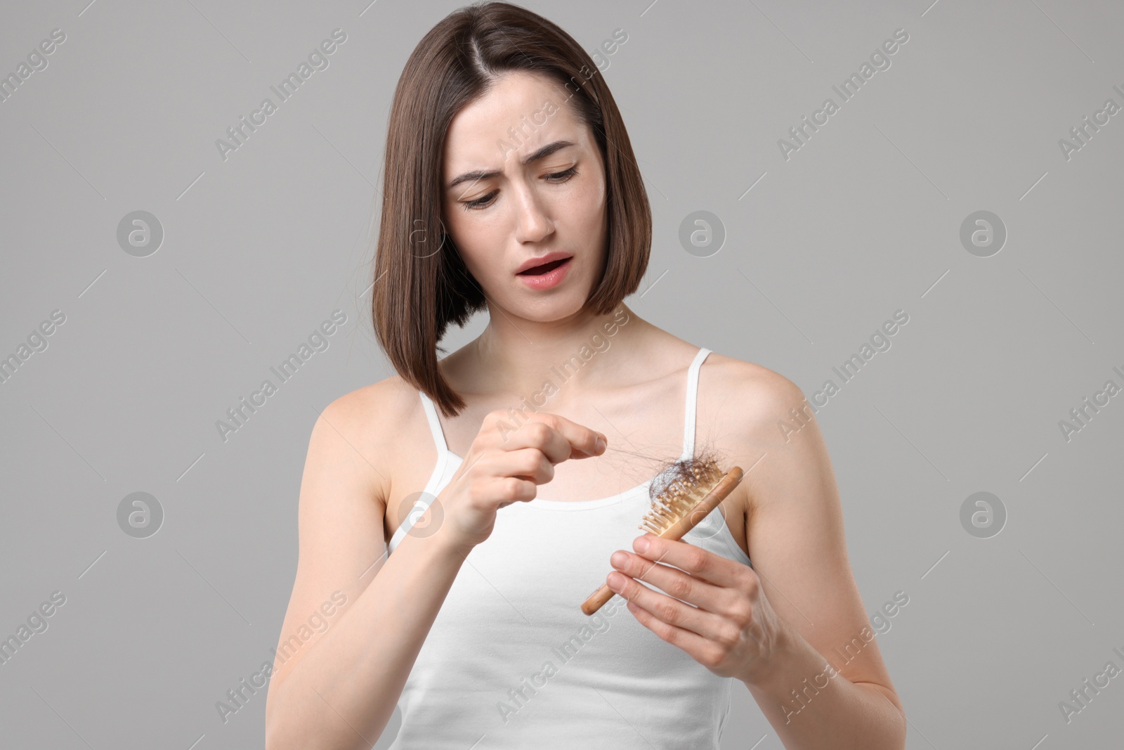 Photo of Stressed woman taking her lost hair from brush on grey background. Alopecia problem