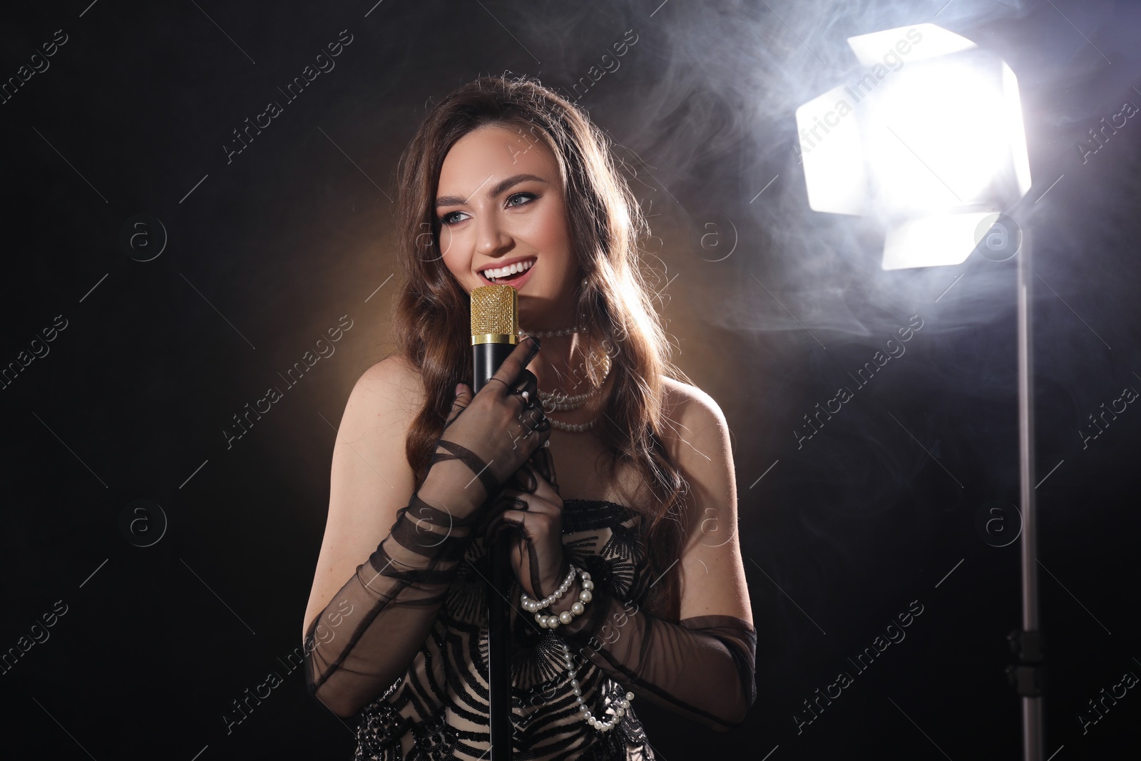 Photo of Beautiful young woman with microphone singing on dark background with smoke