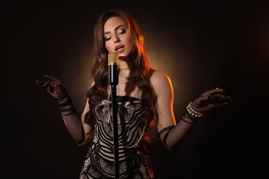 Beautiful young woman in stylish dress with microphone singing on dark background