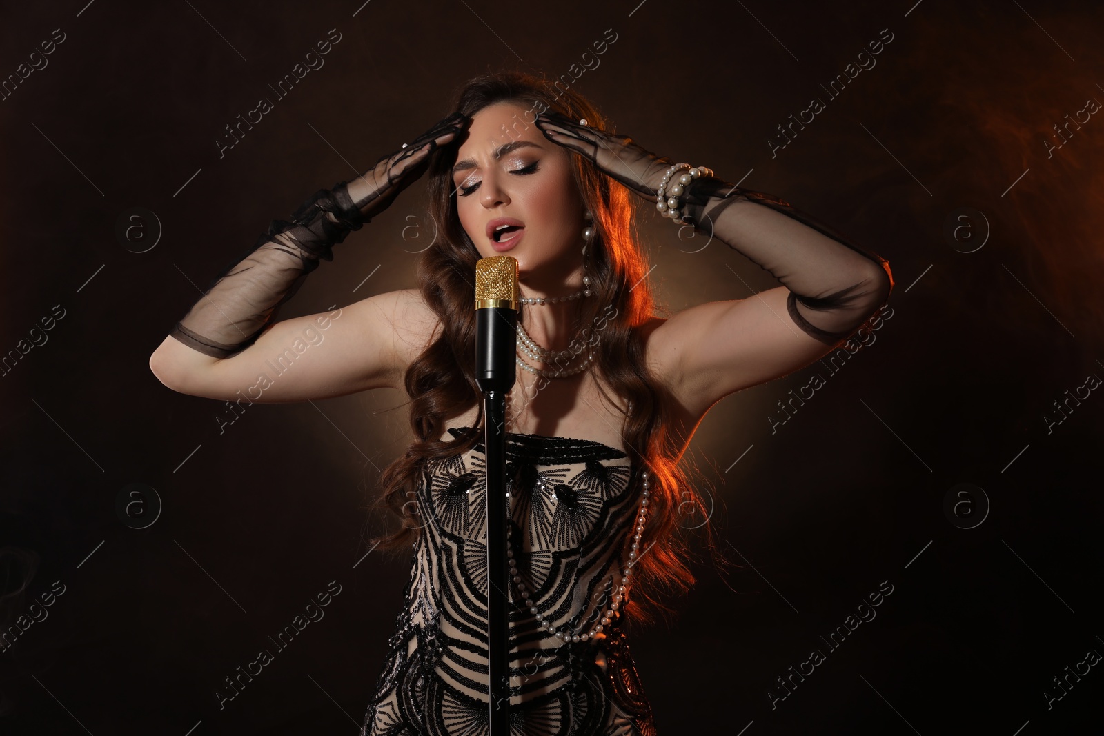 Photo of Beautiful young woman in stylish dress with microphone singing on dark background with smoke