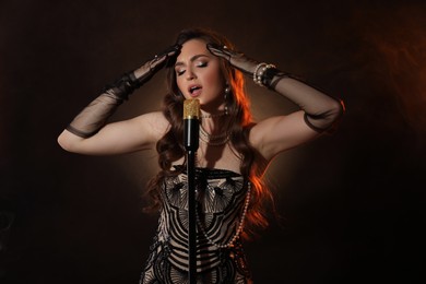 Beautiful young woman in stylish dress with microphone singing on dark background with smoke