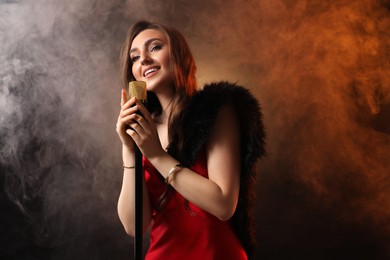 Beautiful young woman in stylish dress with microphone singing on dark background in color lights and smoke