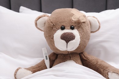 Photo of Toy cute bear with sticking plaster and thermometer under blanket in bed