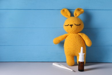 Toy bunny with nasal spray and thermometer on grey table near light blue wooden wall, space for text