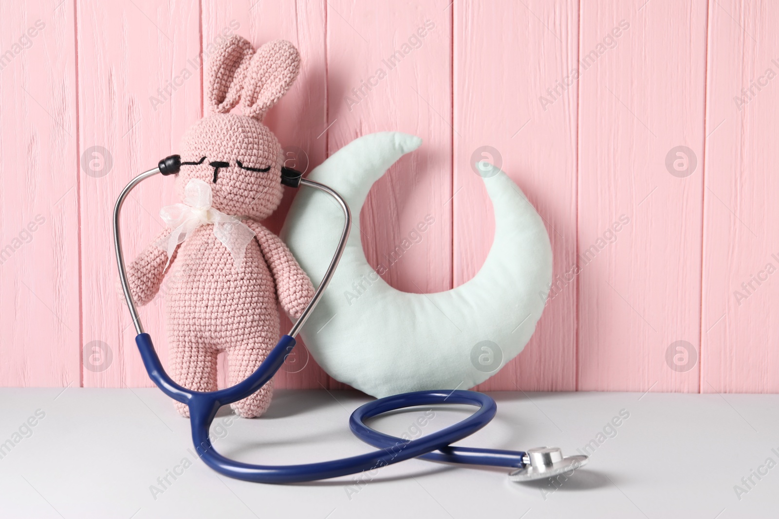 Photo of Toy bunny with stethoscope and pillow on light table near pink wooden wall