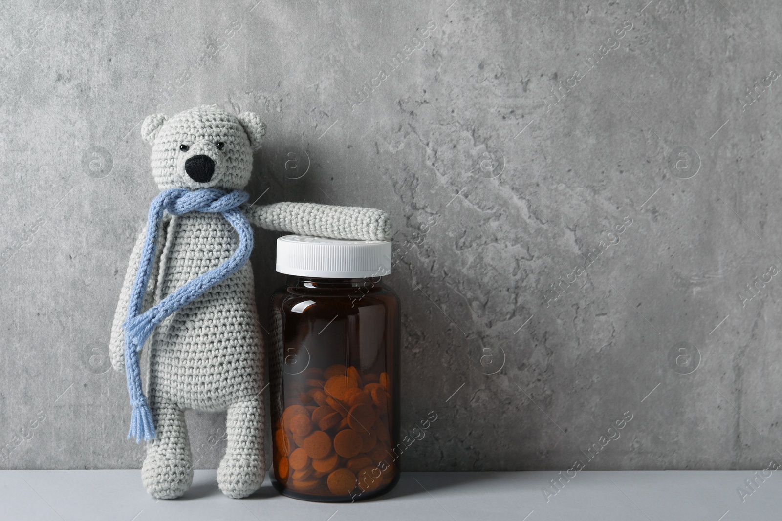 Photo of Toy bear and bottle of pills on light table near grey wall, space for text