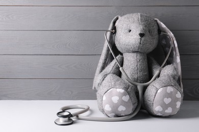 Toy bunny with stethoscope on light table near grey wooden wall, space for text