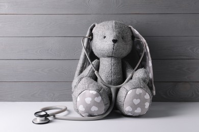 Photo of Toy bunny with stethoscope on light table near grey wooden wall