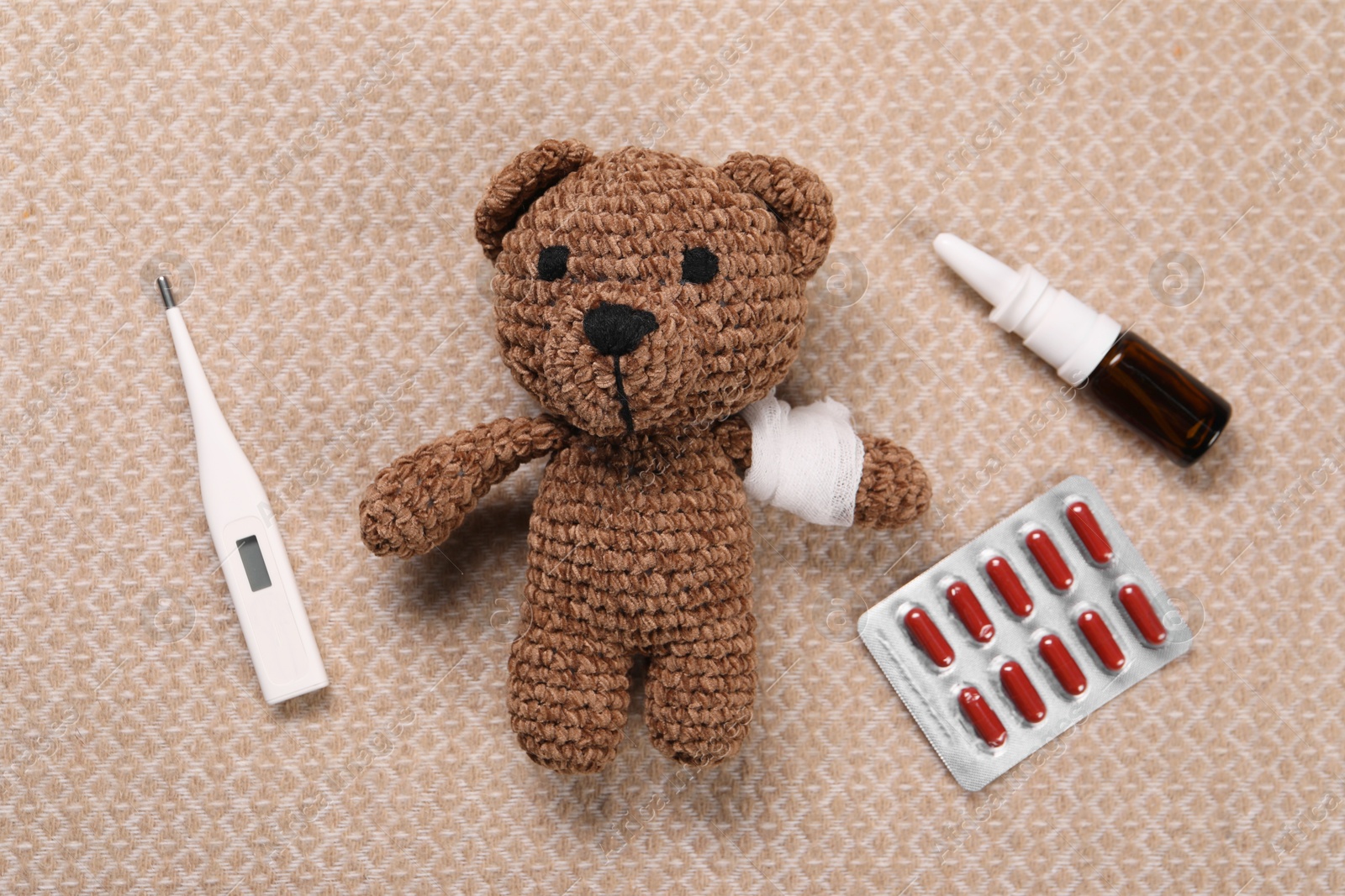 Photo of Toy bear with bandage, thermometer, pills and nasal spray on blanket, flat lay