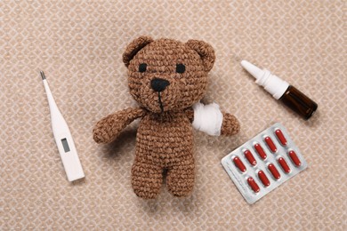 Toy bear with bandage, thermometer, pills and nasal spray on blanket, flat lay