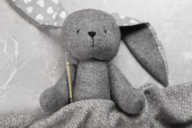 Toy bunny with thermometer under blanket on gray marble background, top view