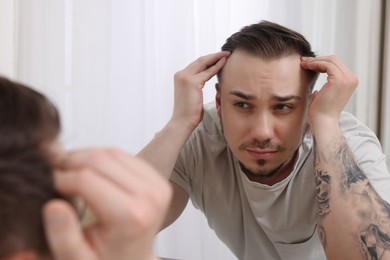 Photo of Baldness concept. Sad man with receding hairline looking at mirror indoors