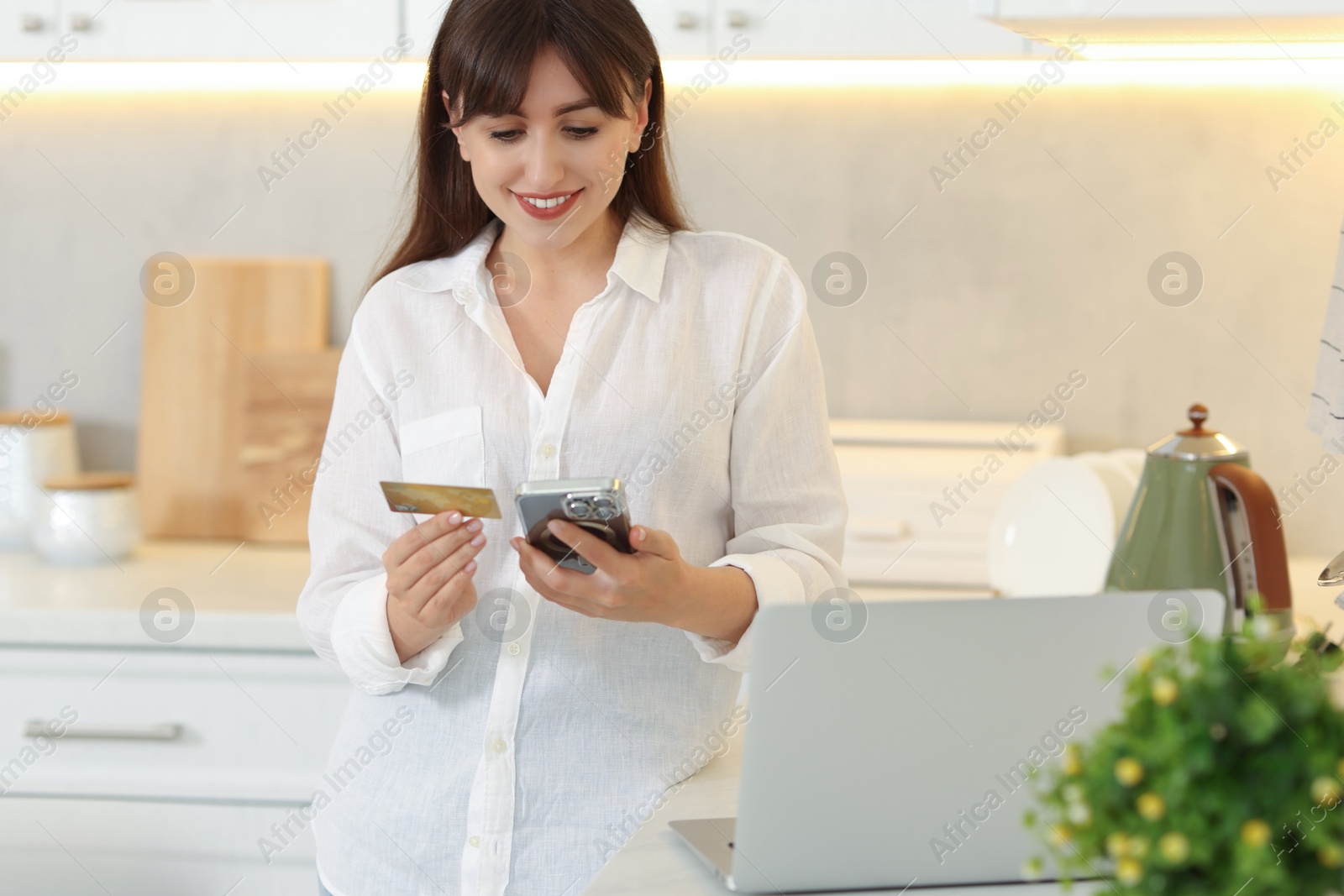 Photo of Online banking. Smiling woman with credit card and smartphone paying purchase in kitchen