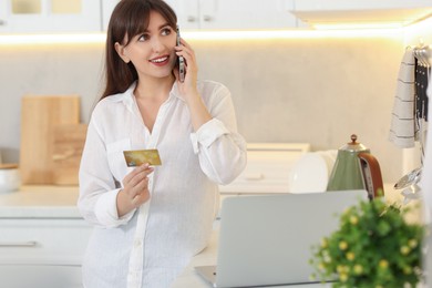 Online banking. Smiling woman with credit card talking by smartphone in kitchen