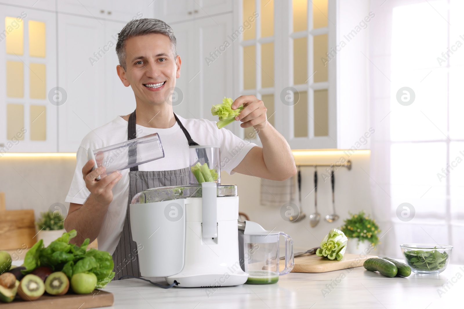 Photo of Smiling man putting fresh celery into juicer at white marble table in kitchen