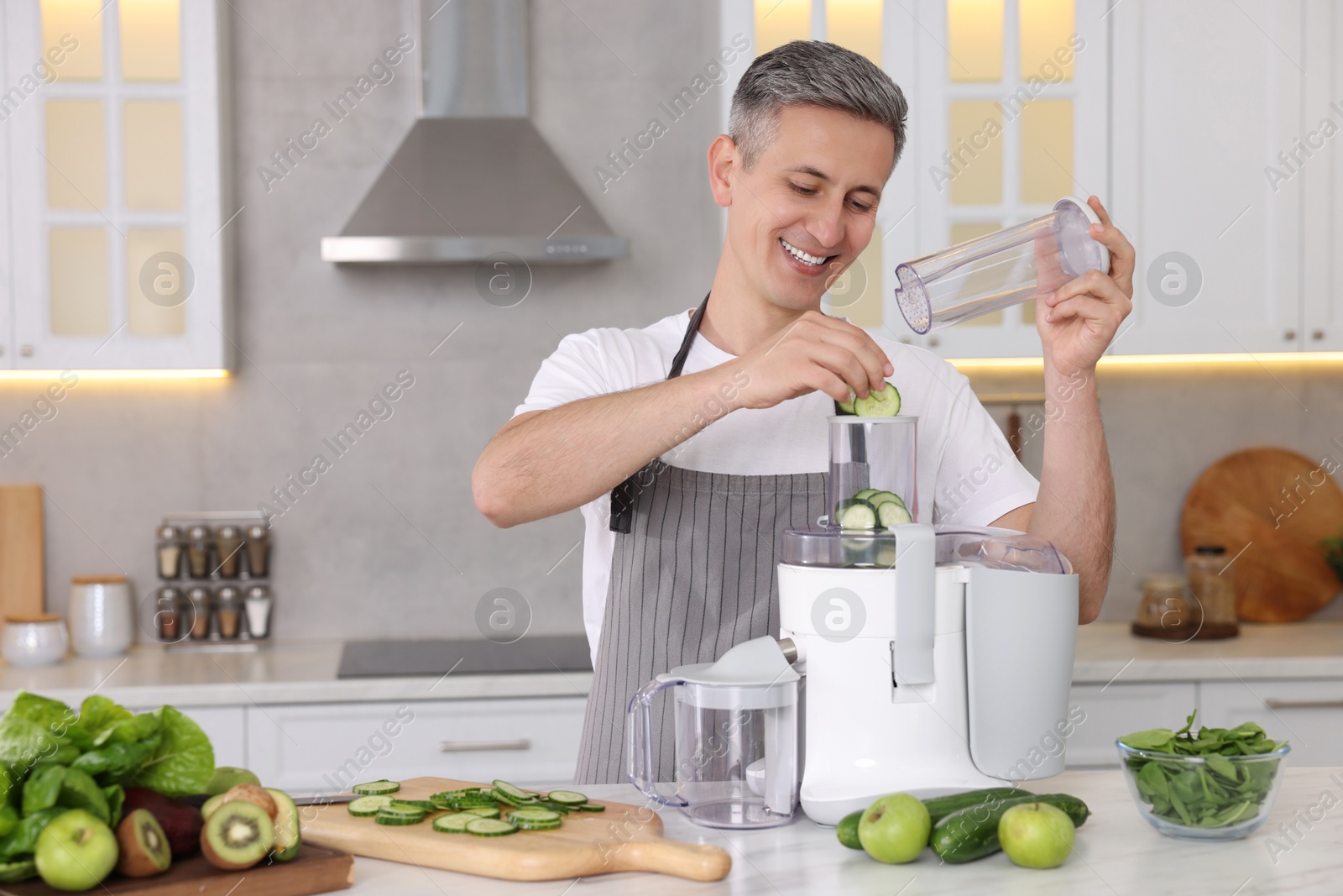 Photo of Smiling man putting fresh cucumber into juicer at white marble table in kitchen