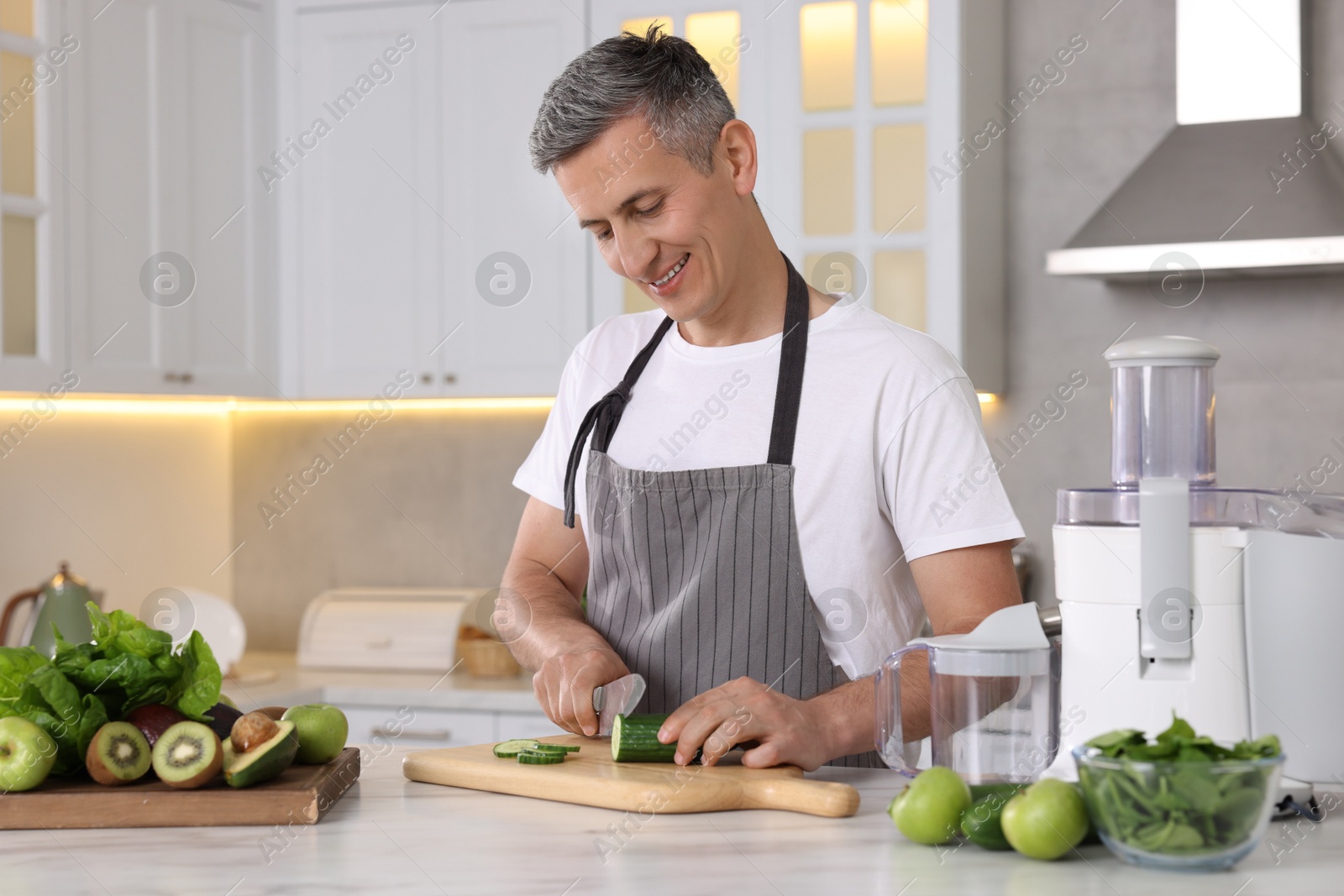 Photo of Juicer and fresh products on white marble table. Smiling man cutting cucumber in kitchen