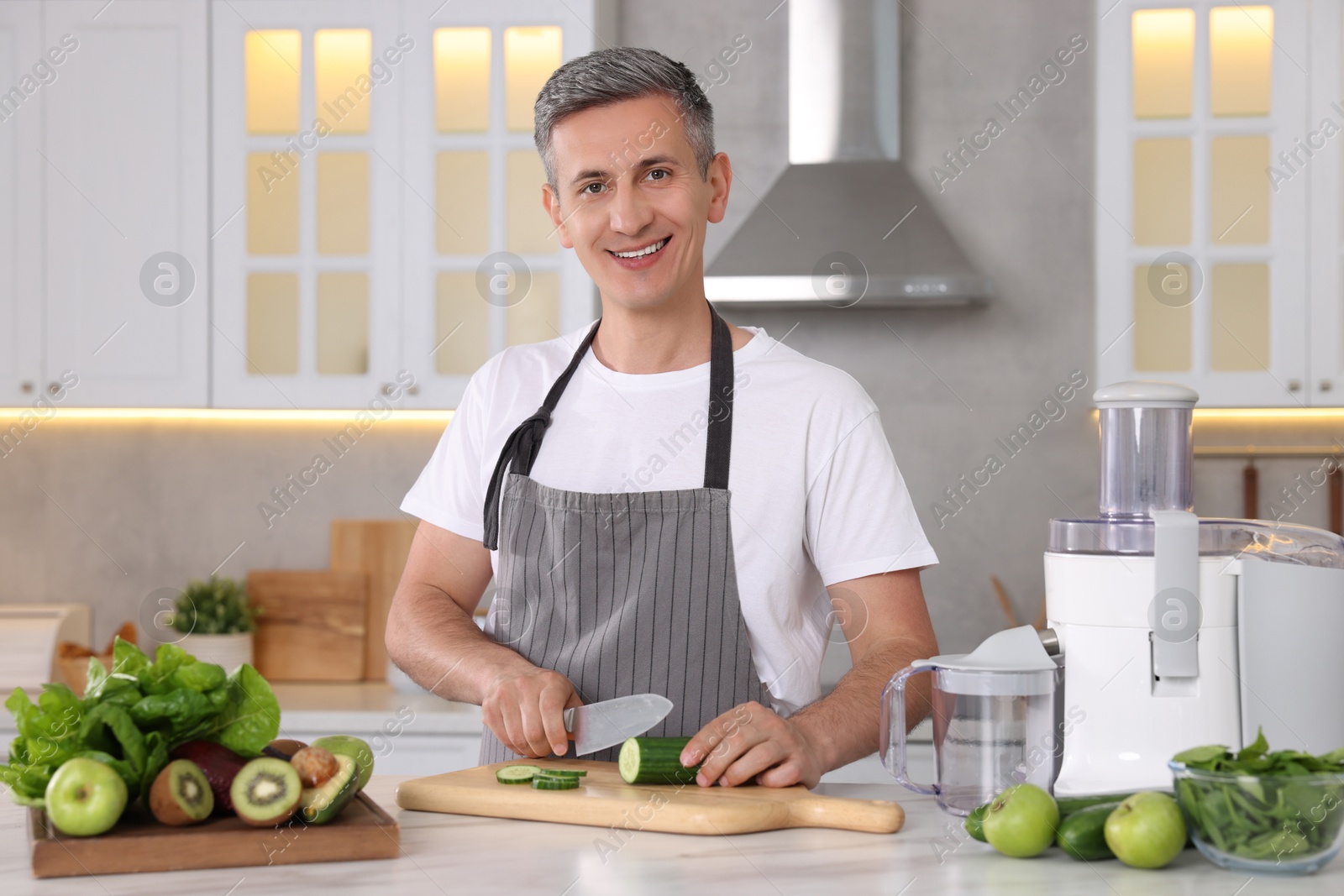 Photo of Juicer and fresh products on white marble table. Smiling man cutting cucumber in kitchen