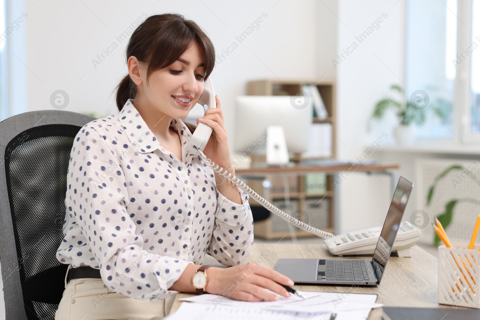 Photo of Smiling secretary talking on telephone at table in office