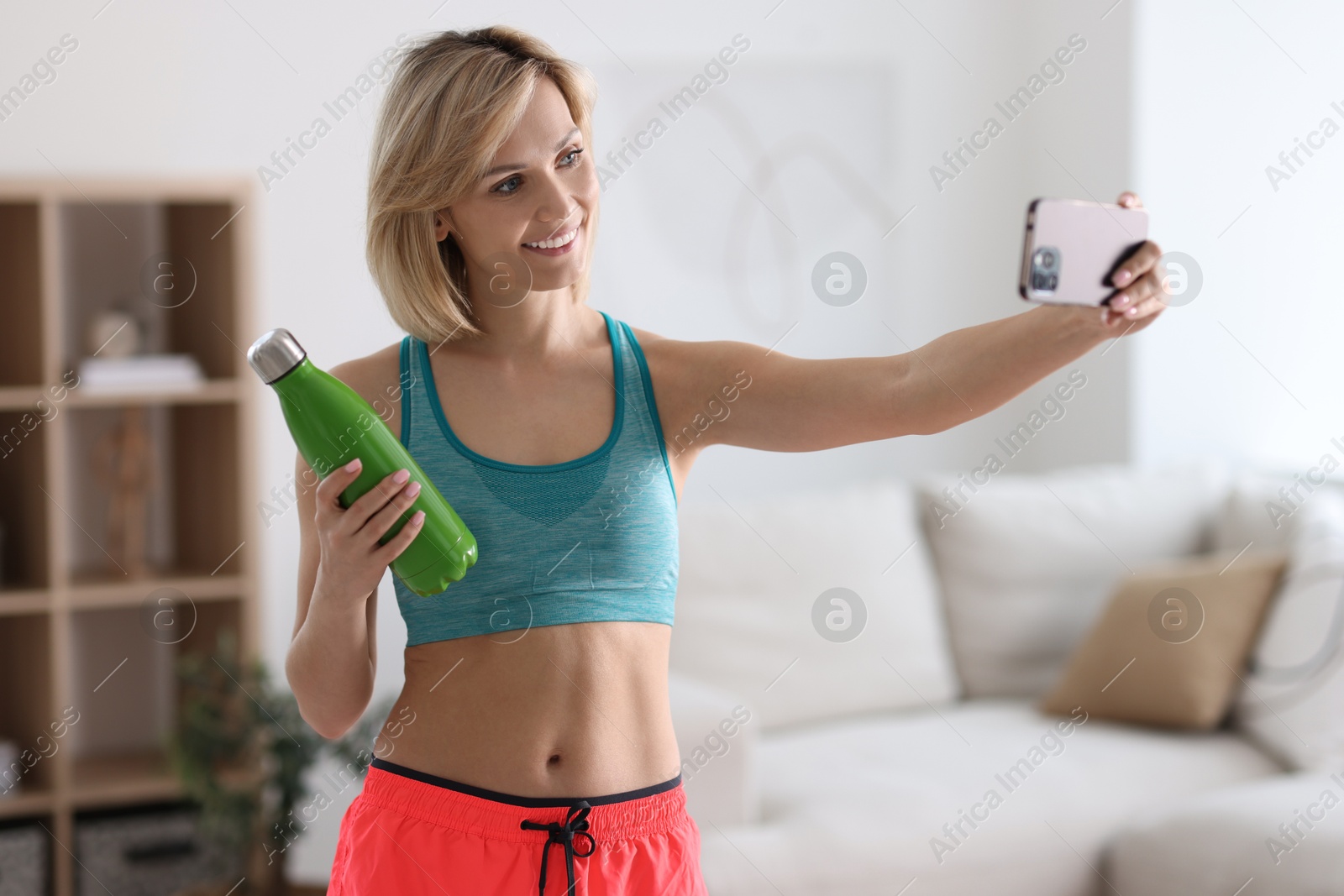 Photo of Smiling sports blogger with bottle streaming online fitness lesson with smartphone at home