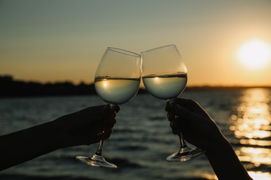 Photo of Women clinking glasses of wine near river at sunset, closeup