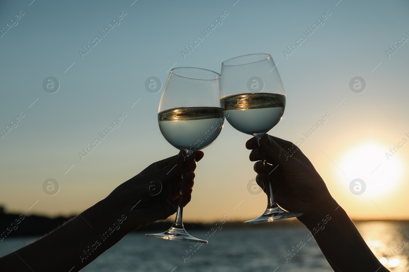 Photo of Women clinking glasses of wine near river at sunset, closeup