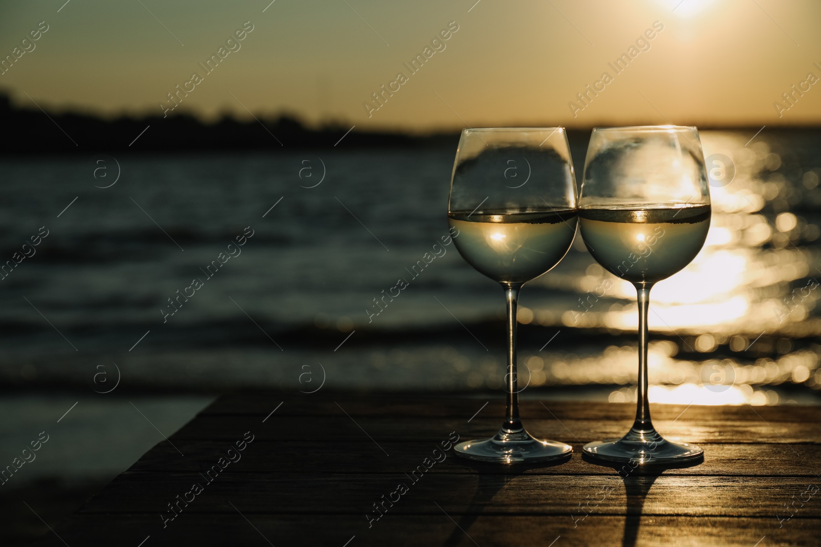 Photo of Two glasses of wine on wooden table near river at sunset. Space for text