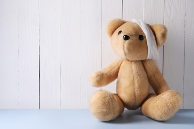Photo of Toy bear with bandage on light blue table near white wooden wall, space for text