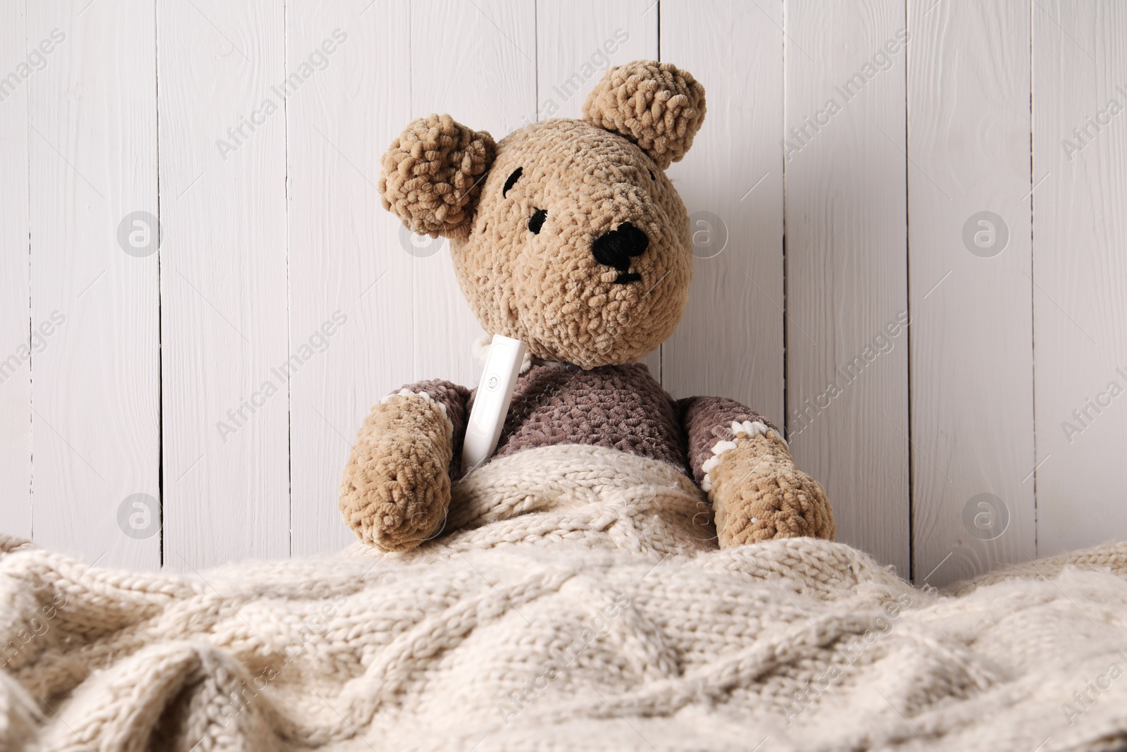 Photo of Toy bear with thermometer under blanket near white wooden wall