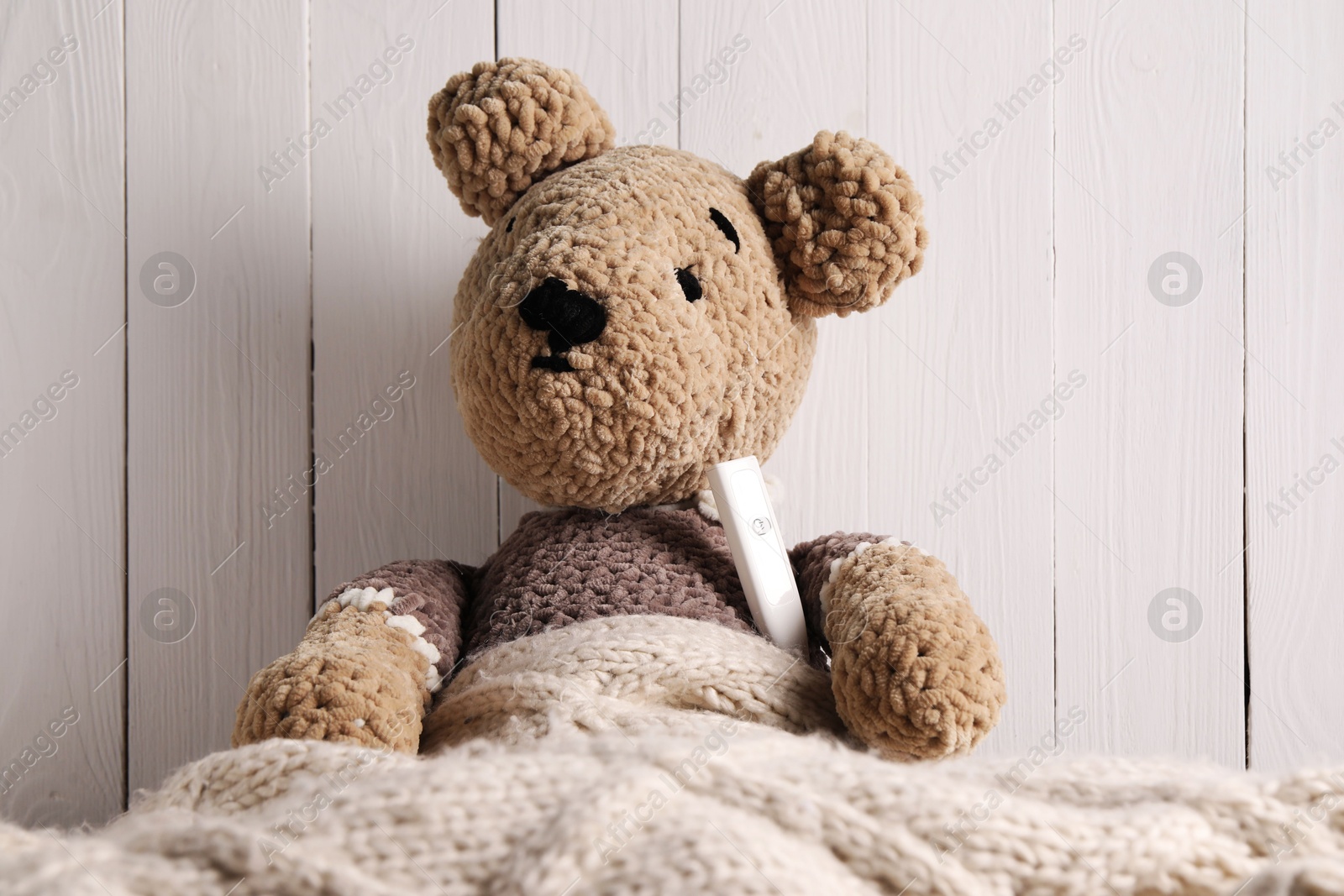 Photo of Toy bear with thermometer under blanket near white wooden wall