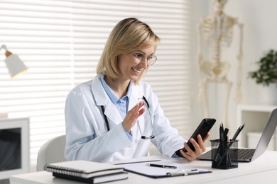 Smiling doctor with smartphone having online consultation at table in office