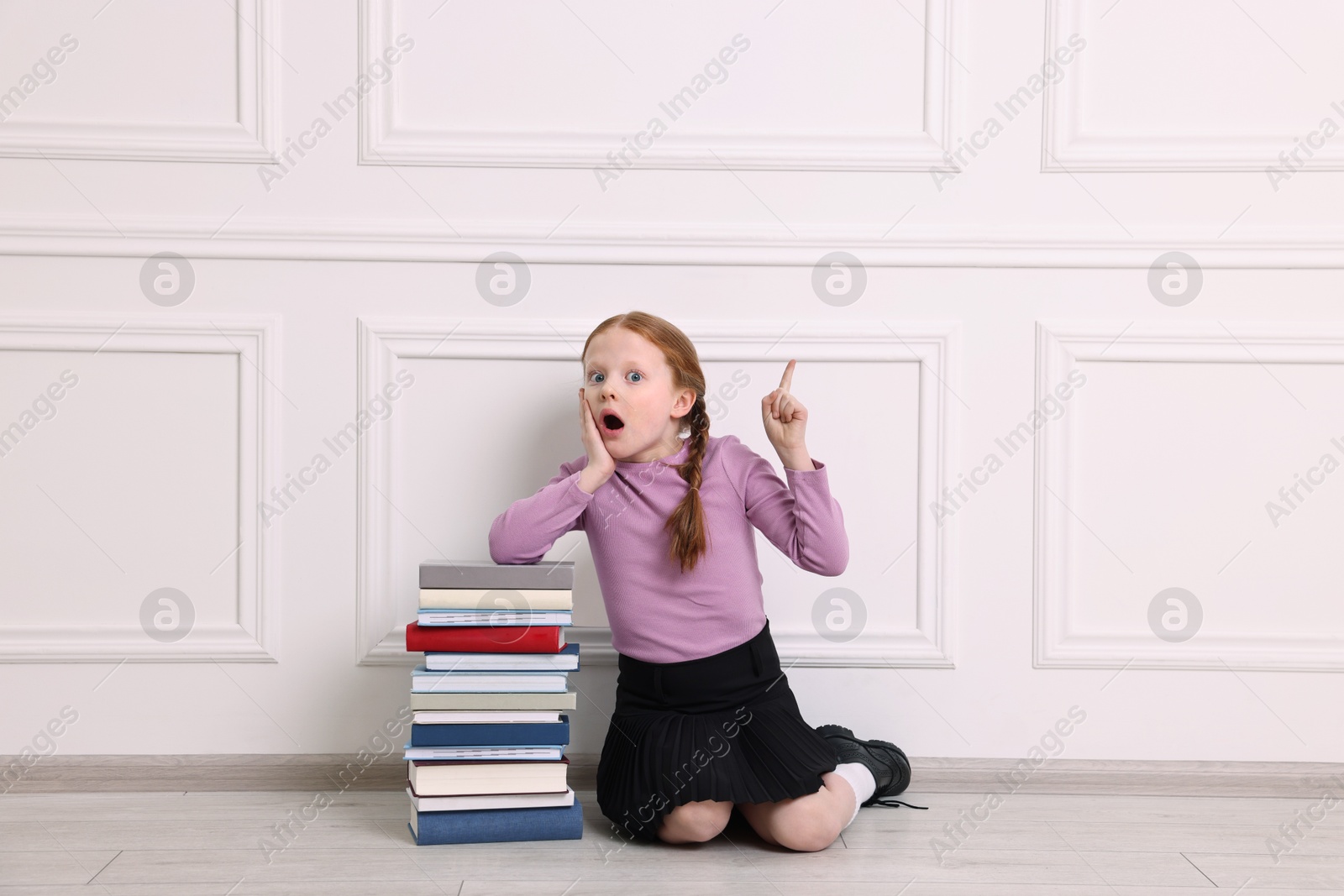 Photo of Surprised girl with stack of books pointing at something indoors