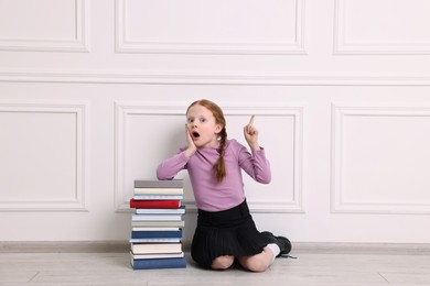 Surprised girl with stack of books pointing at something indoors