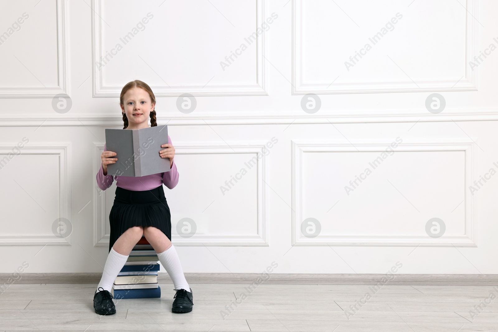 Photo of Little girl sitting on stack of books indoors. Space for text
