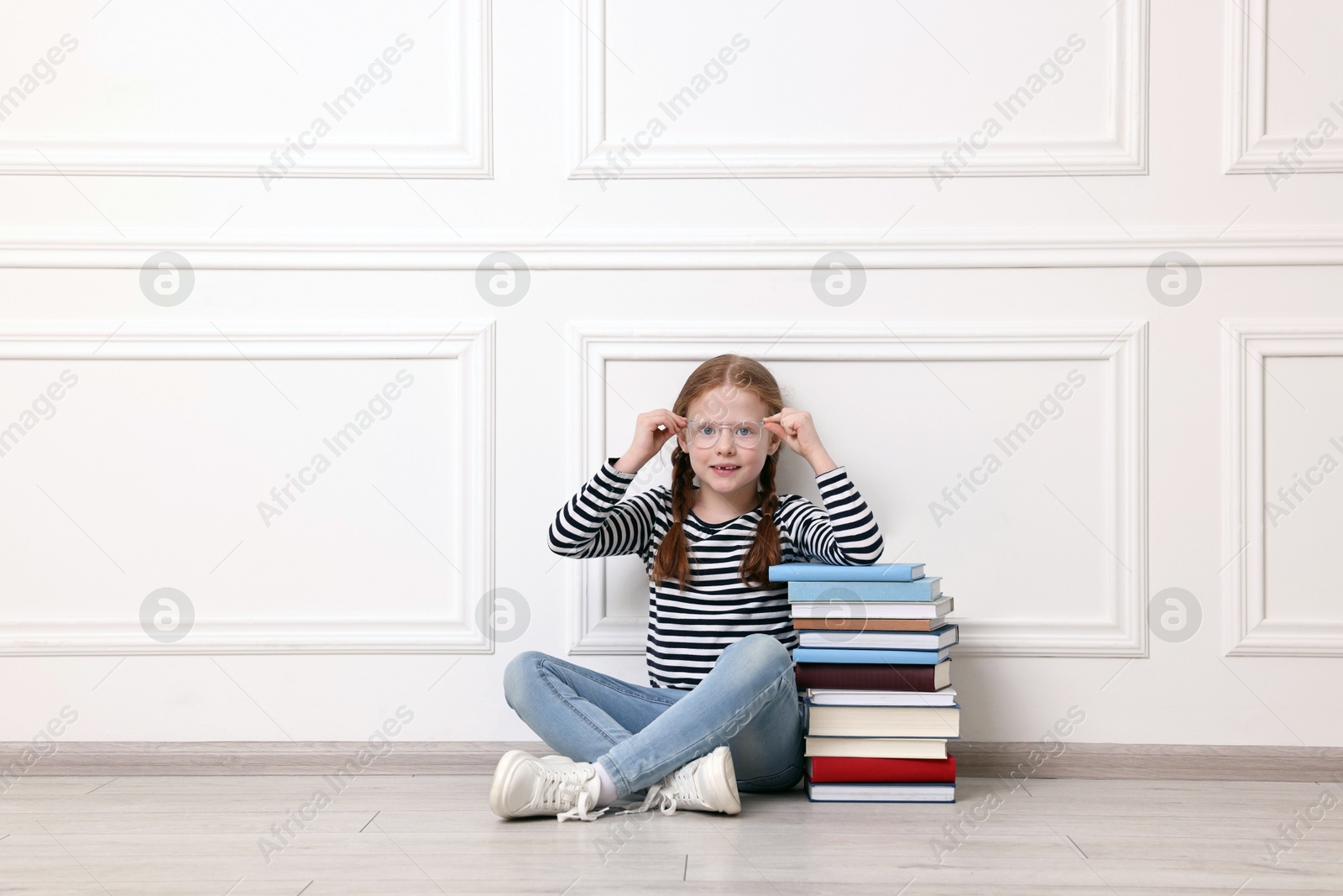 Photo of Cute girl sitting with stack of books indoors