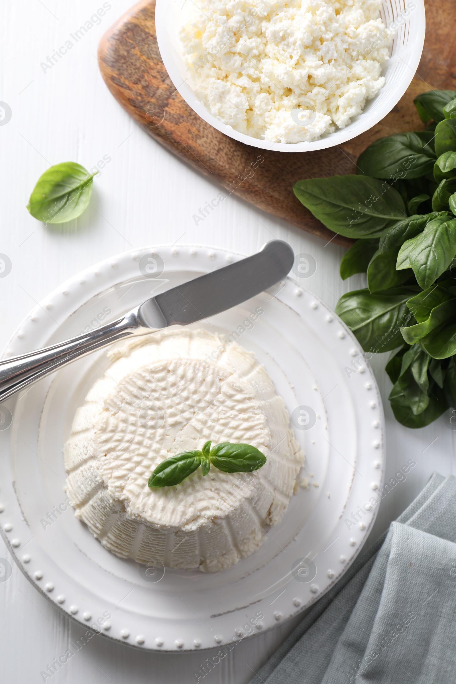 Photo of Fresh ricotta (cream cheese) with basil and knife on white wooden table, flat lay